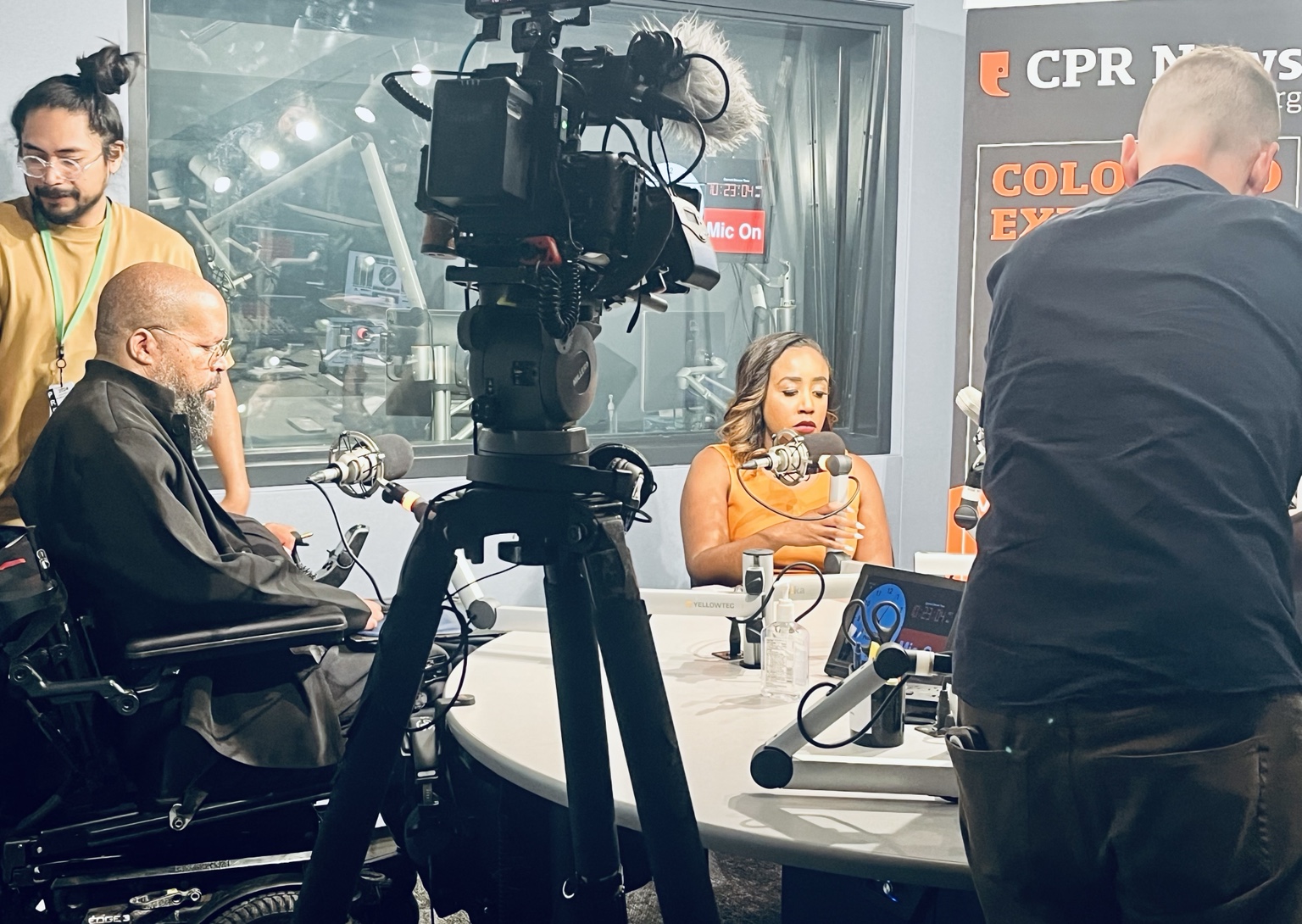 Stewart Tucker Lundy at a microphone in the CPR News studio, with a camera crew and another speaker.